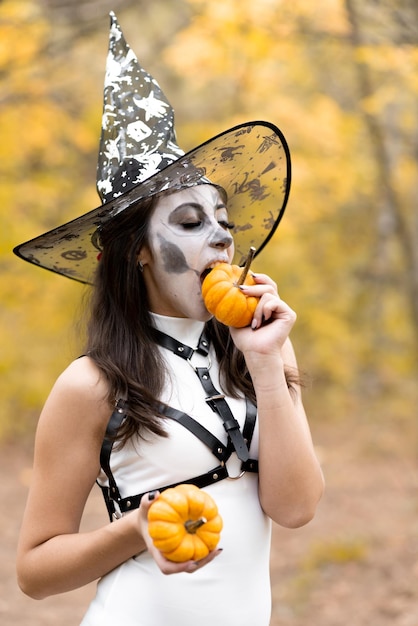 Halloween Young beautiful girl in a white dress with makeup on her face in a witch39s hat posing in the autumn forest Skeleton face makeup Bites a small pumpkin The day of the Dead