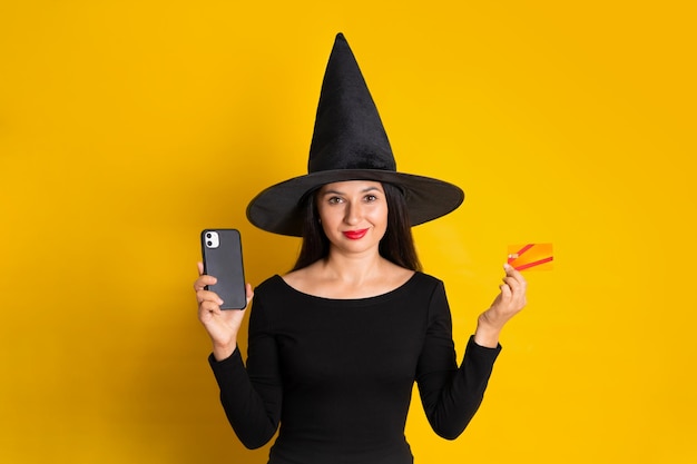 Halloween A young beautiful woman in a witch costume holds a phone and a bank card in her hands Sale Discount Yellow background space for text