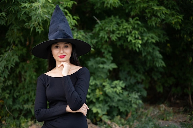 Halloween A young beautiful woman in a witch39s hat and a black dress in the park Space for text