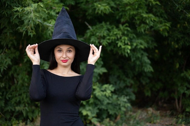 Halloween A young beautiful woman in a witch39s hat and a black dress in the park Space for text