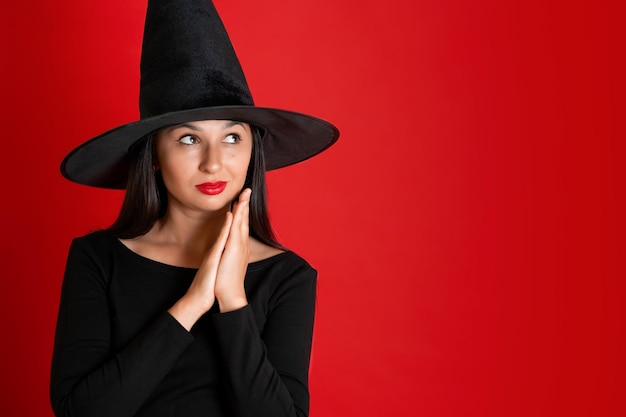 Halloween A young beautiful woman in a witch39s hat and a black dress on a red colored background Space for text