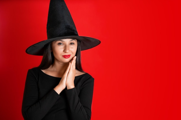 Halloween A young beautiful woman in a witch39s hat and a black dress on a red colored background Space for text