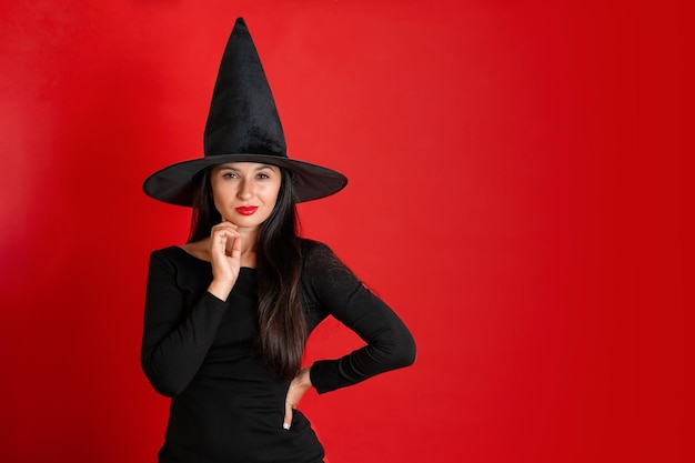 Halloween A young beautiful woman in a witch39s hat and a black dress on a red colored background Space for text