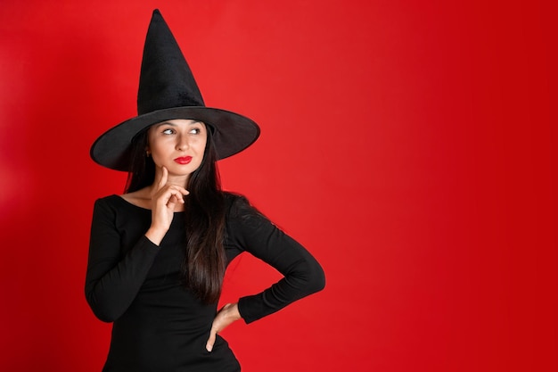 Halloween A young beautiful woman in a witch39s hat and a black dress on a red colored background Space for text