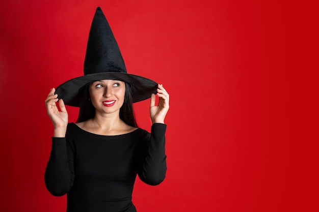 Halloween A young beautiful woman in a witch39s hat and a black dress on a red colored background Space for text