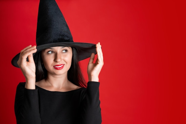 Halloween A young beautiful woman in a witch39s hat and a black dress on a red colored background Space for text