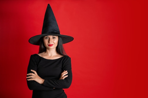 Halloween A young beautiful woman in a witch39s hat and a black dress on a red colored background Space for text