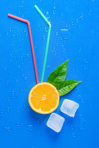 Halved Orange, Green Leaves, Drinking Straws and Ice Cubes on Blue Background.