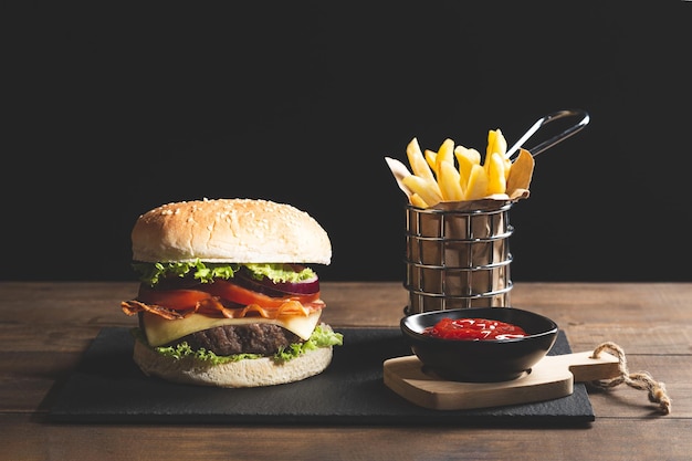 Hamburger chips and ketchup on wooden base on dark background