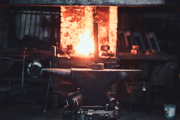 Hammer on anvil at dark blacksmith workshop with fire in stove at background.