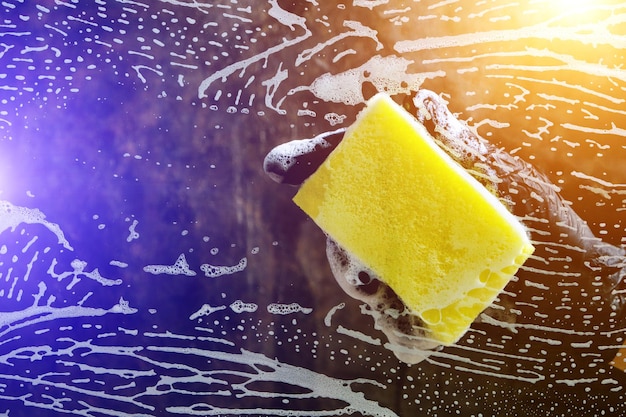 Hand in a black glove washes the glass with a foam sponge with foam on a dark background with light.