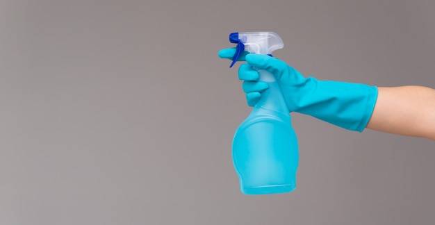 A hand in blue rubber glove holds the glass cleaner in a spray bottle on a neutral background.