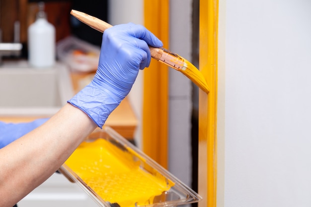 Hand in blue rubber glove with paintbrush painting natural wooden door with yellow paint