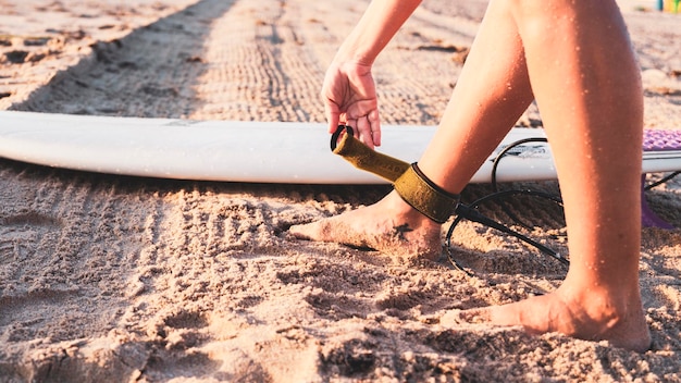 Photo a hand buckling the leash on the ankles of a girl on the sandy beach with a surfboard