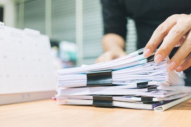 Hand of businessman for searching information in Stacks of paper on work desk office.