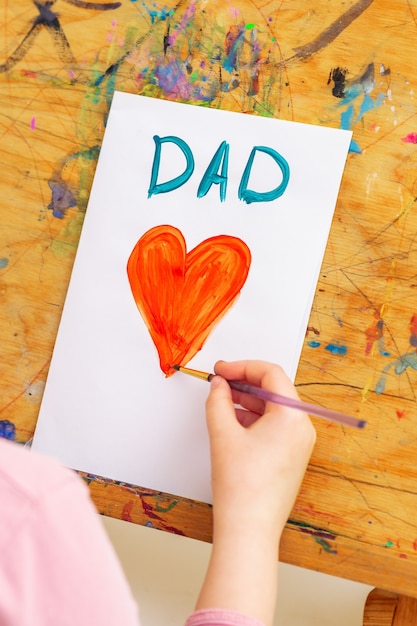 Photo hand of child drawing red heart with word dad greeting card on white paper on an easel. family and father's day concept.