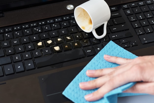 Hand cleans spilled coffee on laptop keyboard with rag