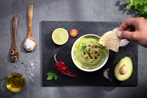 Hand dipping nachos in guacamole on dark background. Top view.