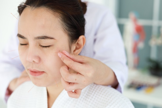 Hand of doctor performing acupuncture therapy . Asian female undergoing acupuncture treatment with a line of fine needles inserted into the her face skin in clinic hospital