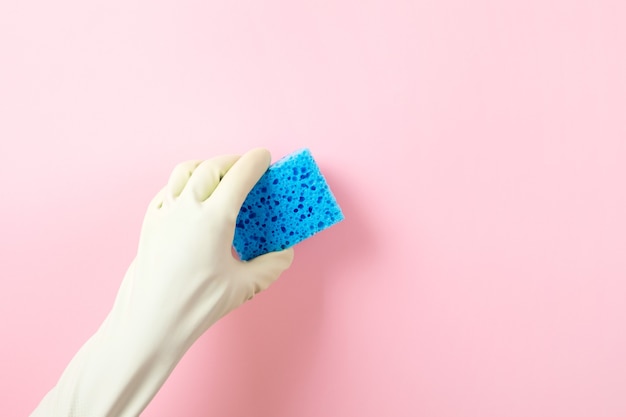 Hand in a green rubber glove holds a blue  washing sponge over pink background