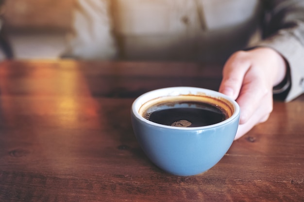 hand holding a blue cup of hot black coffee on wooden table in cafe