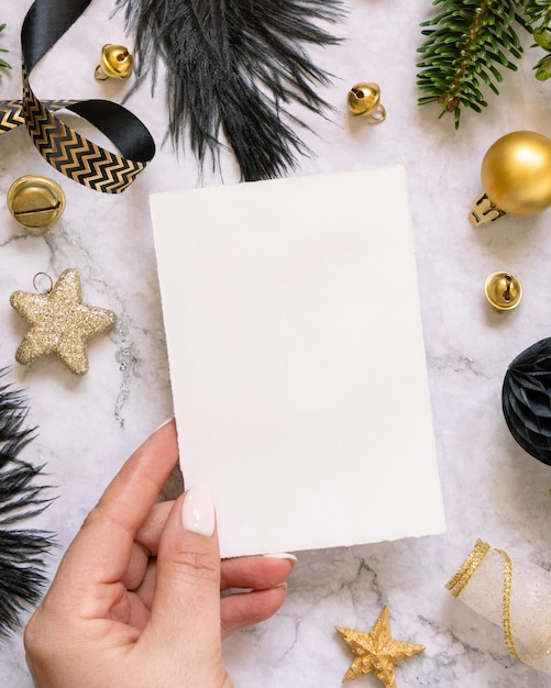 Hand holding Card near black and golden Christmas decorations and fir branches Mockup