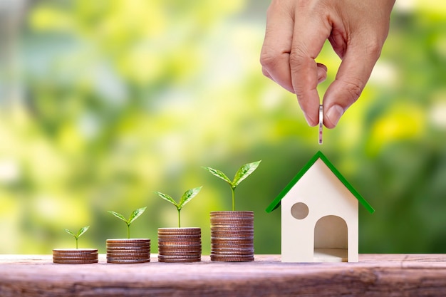 Hand holding coins put in house model for saving money and trees growing on a pile of money finance