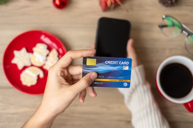 Hand holding credit card for online shopping on smartphone during eating Homemade Xmas Cookies and coffee. Merry Christmas eve, December, seasonal, Black Friday sale, New Year and holiday concept