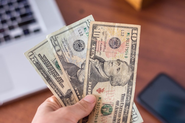 Hand holding dollars bills with smartphone and laptop in background