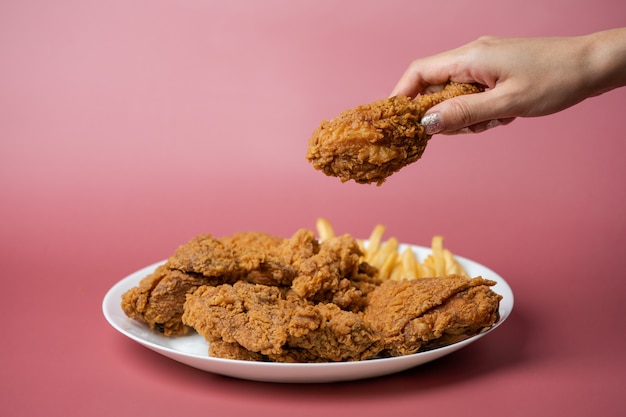 Hand holding drumsticks, crispy fried chicken with french fries in white plate  on red background.