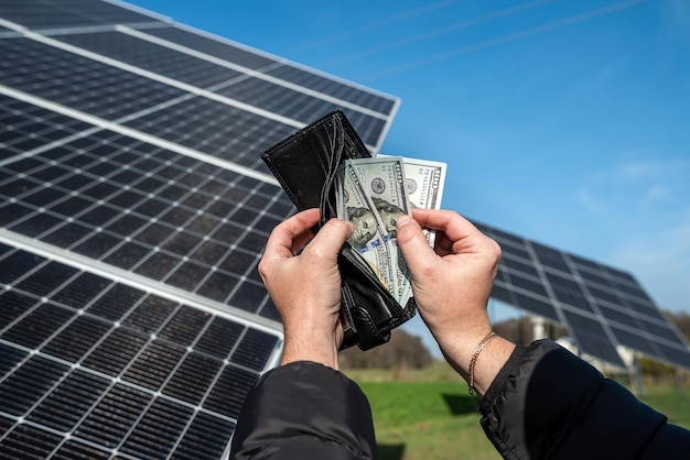 A hand holding a fan of dollars in a wallet near solar panels US dollar banknotes on the background The concept of paying for solar panels