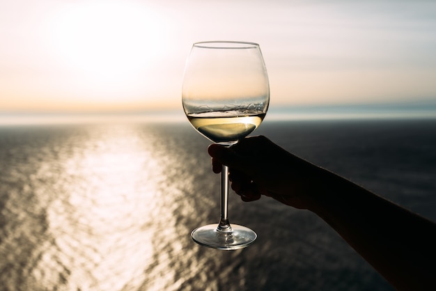 Photo hand holding glass of wine over the sea. romantic vacation. hand holding a glass of wine against the sea. meeting the sunset with wine by the sea. copy space