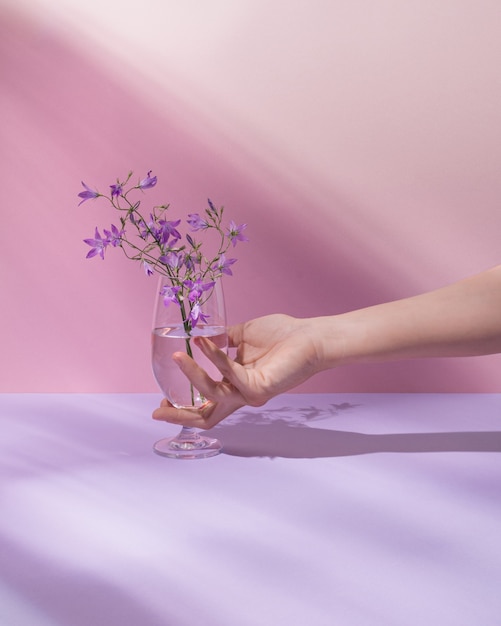 Hand holding glass with transparent liquid and beautiful purple field flowers. Pastel pink summer or spring background. Minimal nature, aesthetic. Natural sun rays and shadows.