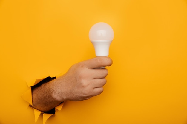 Hand holding an incandescent led light bulb from a torn hole in yellow paper