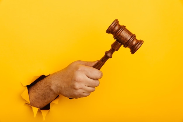 Hand holding a judge's gavel through torn yellow background Law and auction aconcept