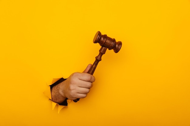 Hand holding a judge's gavel through torn yellow paper wall Law concept