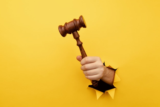 Hand holding a judges gavel through torn yellow paper wall law and justice concept