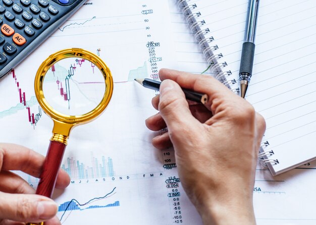 Hand holding a magnifying glass and pencil above economic charts