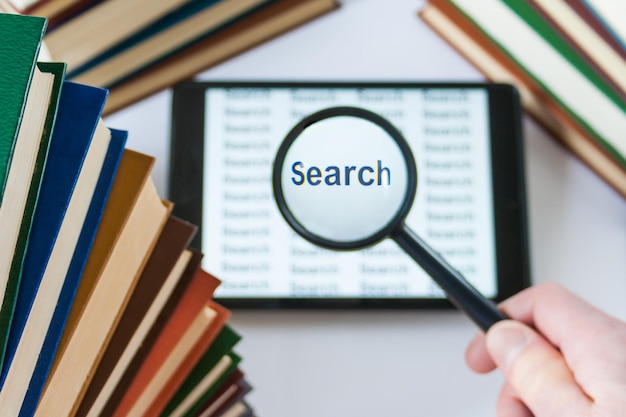 Photo hand holding a magnifying glass over a tablet surrounded by books search concept