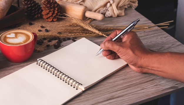 Hand holding pen is writting white notebook spread on a table with coffee and bread placed.