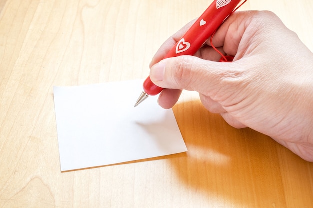 Hand holding red pen with white heart pattern, writing on blank notepad paper on wooden table