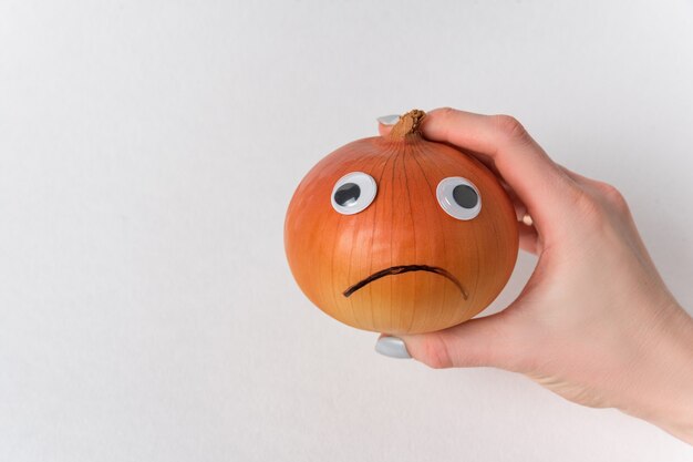 Hand holding sad onion on white background. Napiform onion with funny face and Googly eyes.