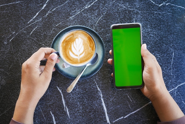 hand holding a smart phone with green screen and cup of coffee on table