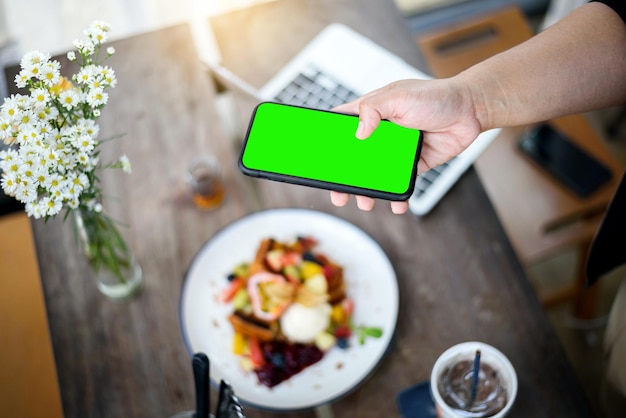 Hand holding smartphone taking French toast topping mixed fruit ice cream italian soda on wooden table in modern bright kitchen interior green screen