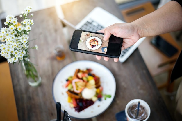 Hand holding smartphone taking French toast topping mixed fruit ice cream italian soda on wooden table in modern bright kitchen interior