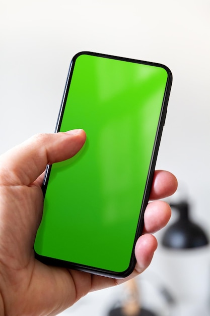 Hand holding a smartphone with blank green screen Office background