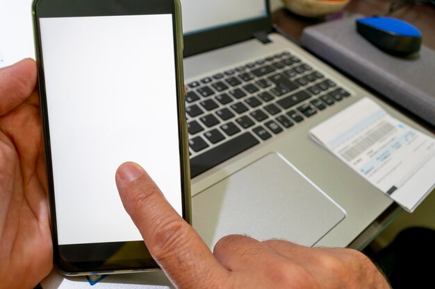 Hand holding smartphone with finger on white screen and computer in the background.