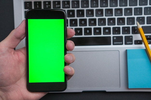 Hand holding Smartphone with Green Screen Mock Up Display. In the background is a laptop