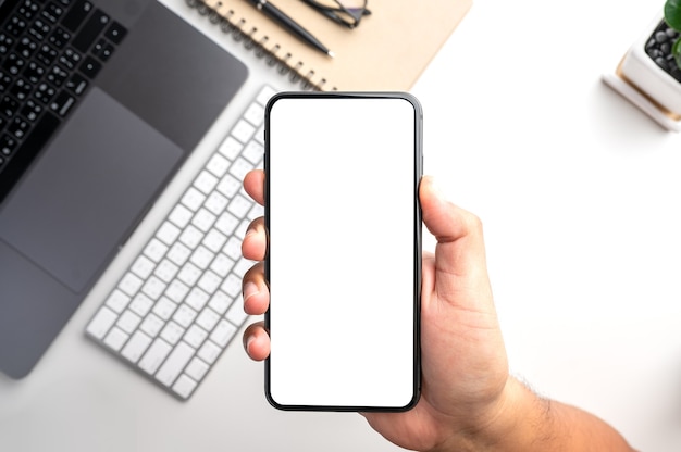 hand holding smartphone with white blank screen over desktop with laptop computer