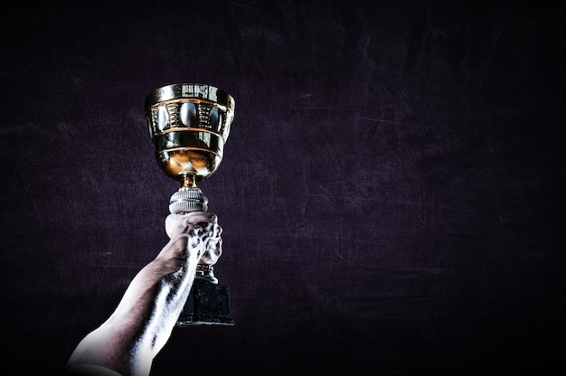 Hand holding up a gold trophy cup against dark background. Mixed media